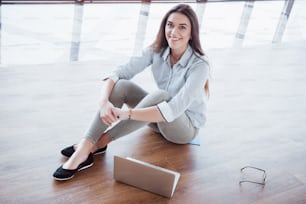 Side view of attractive girl using a laptop in public wifi area and smiling while sitting on the floor.