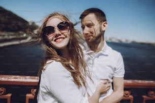 happy woman with windy hair in sunglasses smiling, stylish couple in love having fun on bridge in the summer city. modern woman and man in fashionable white clothes embracing at the river