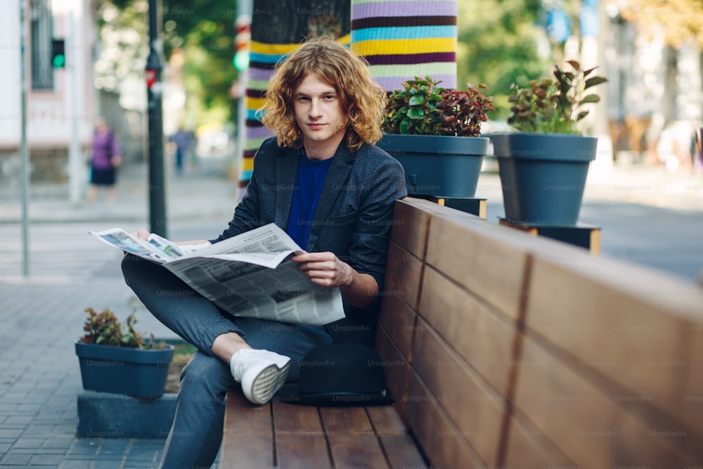 Young smiling reddish, red haired hipster man with curly hair, jacket and T-shirt, holding a newspaper while sitting leg over leg on a long wooden bench in the city outdoors, looking to camera.