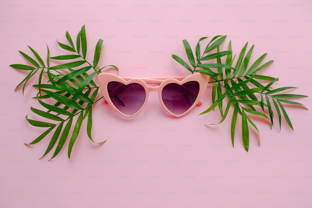 conceito de férias de verão. óculos de sol rosa elegantes em forma de coração e folhas de palmeira verde no fundo rosa, flat lay. espaço para texto.  tempo para viajar conceito e relaxar. Férias