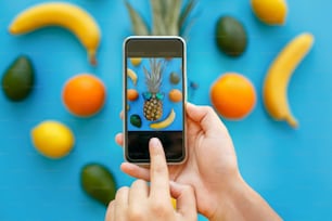 Hände, die das Telefon halten und Fotos von Ananas in Sonnenbrillen und Bananen, Orangen und Avocado auf blauem Papier machen, trendige flache Liege. Stilvolle Food-Fotografie. Instagram Obst Foto