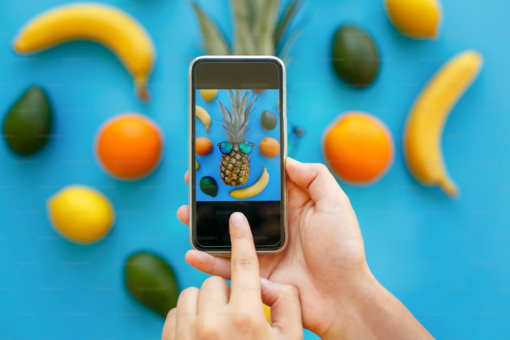 hands holding phone and taking photo of pineapple in sunglasses and bananas, oranges and avocado on blue paper, trendy flat lay. stylish food photography. instagram fruit photo