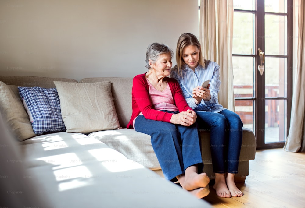 Una abuela anciana y una nieta adulta con un teléfono inteligente en casa, sentadas en un sofá.