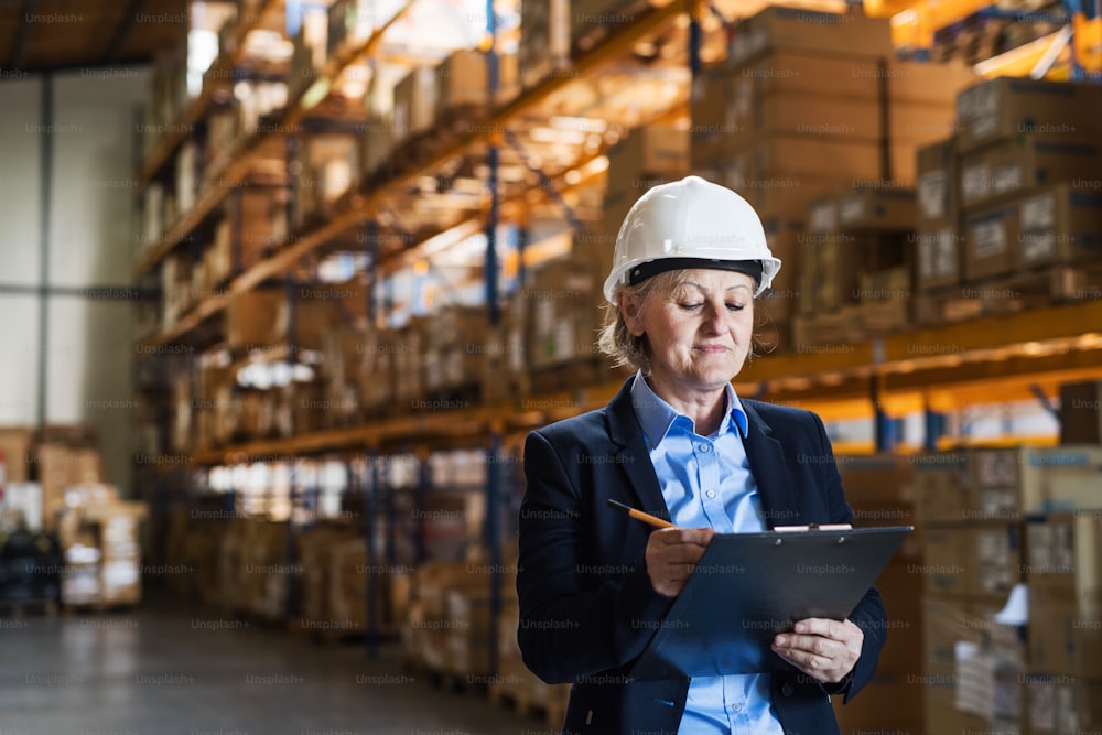 Senior woman warehouse manager or supervisor with a white helmet and clipboard, controlling stock.