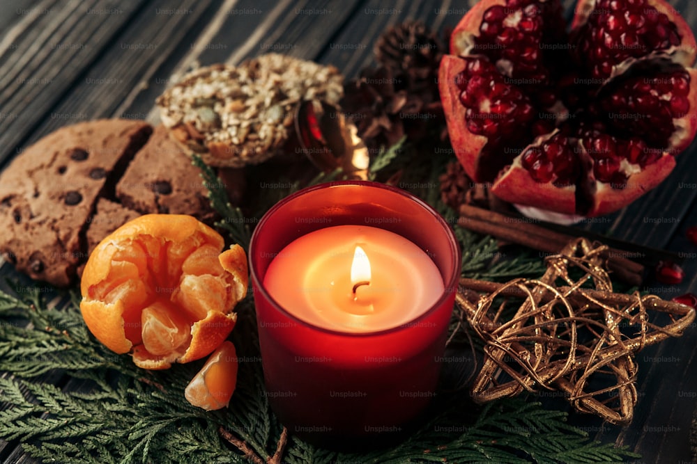 スタイリッシュで素朴なクリスマスの壁紙キャンドルと緑の枝と木製の背景に赤いリボンとクッキーの果物をプレゼントします。冬休みカードのコンセプトの季節の挨拶