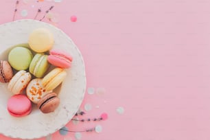 delicious colorful macaroons in vintage white plate on trendy pastel pink paper, stylish flat lay. tasty pink, yellow, green and brown macarons with lavender and confetti. space for text