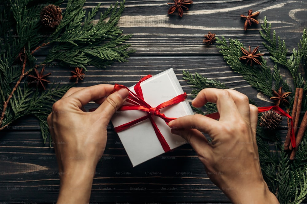 Stilvolle Weihnachtswohnung lag mit Händen, die Geschenk mit rotem Band auf rustikalem Holzhintergrund mit grünen Zweigen umwickeln. Platz für Text. Saisonale Grüße für Winterurlaub Kartenkonzept