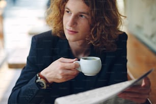 클로즈업 초상화. 곱슬곱슬한 붉은 머리를 가진 젊은 흥미로운 남자, 재킷과 빈티지 시계를 착용하고 신문을 읽고 커피를 마시며 야외 카페에서 왼쪽을 바라보고 있습니다.