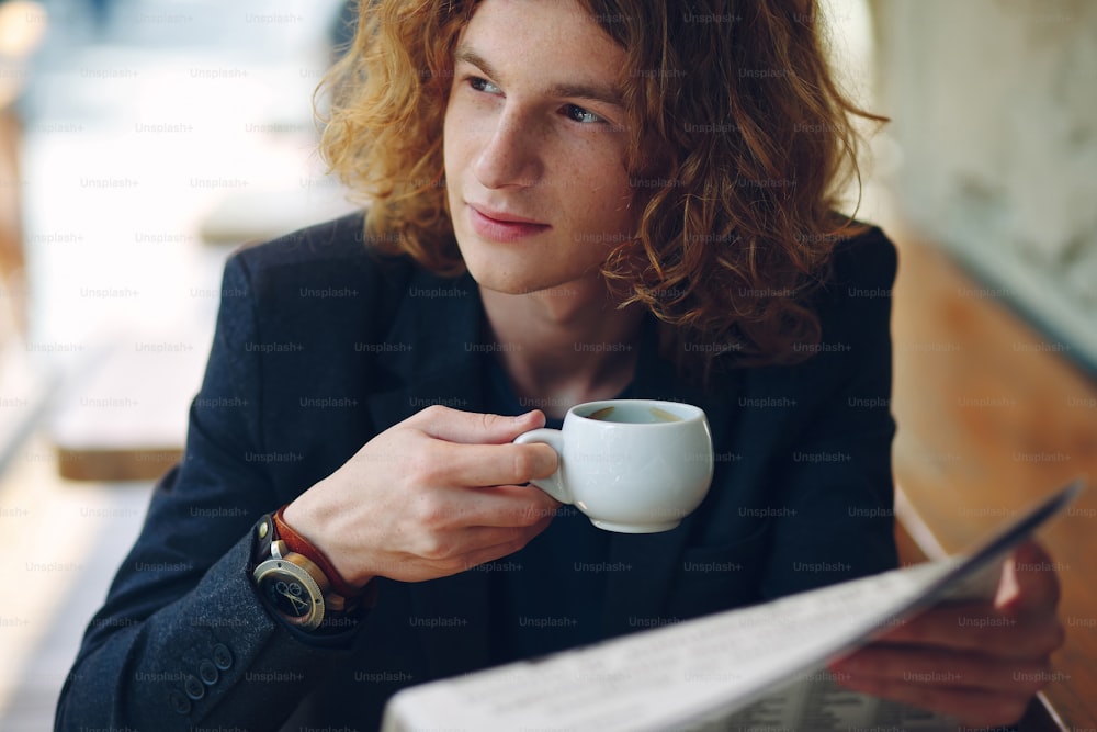 Close-up portrait. Young interesting man with curly reddish hair, wearing jacket and vintage watch, reading newspaper and drinking coffee while looking to left in a cafe outdoor.