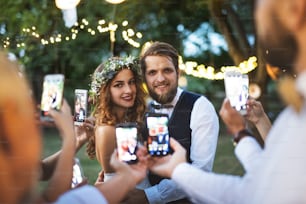 Invitados con teléfonos inteligentes tomando fotos de la novia y el novio en la recepción de la boda afuera en el patio trasero.