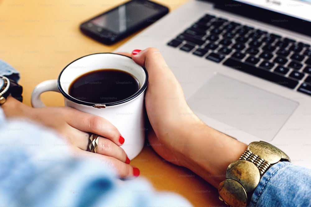 Giovane ragazza elegante che tiene una tazza di caffè su sfondo artigianale con laptop e telefono, concetto di casa freelance e di lavoro