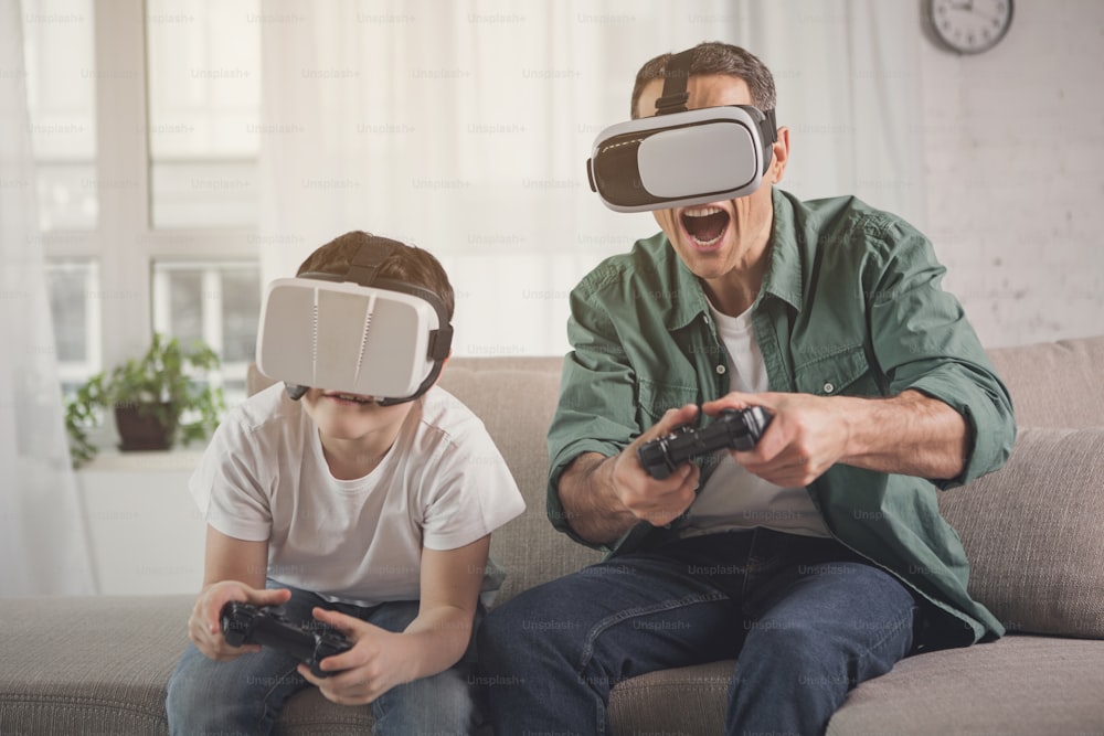 Cheerful family enjoying video game competition. They are holding joysticks and wearing virtual reality googles