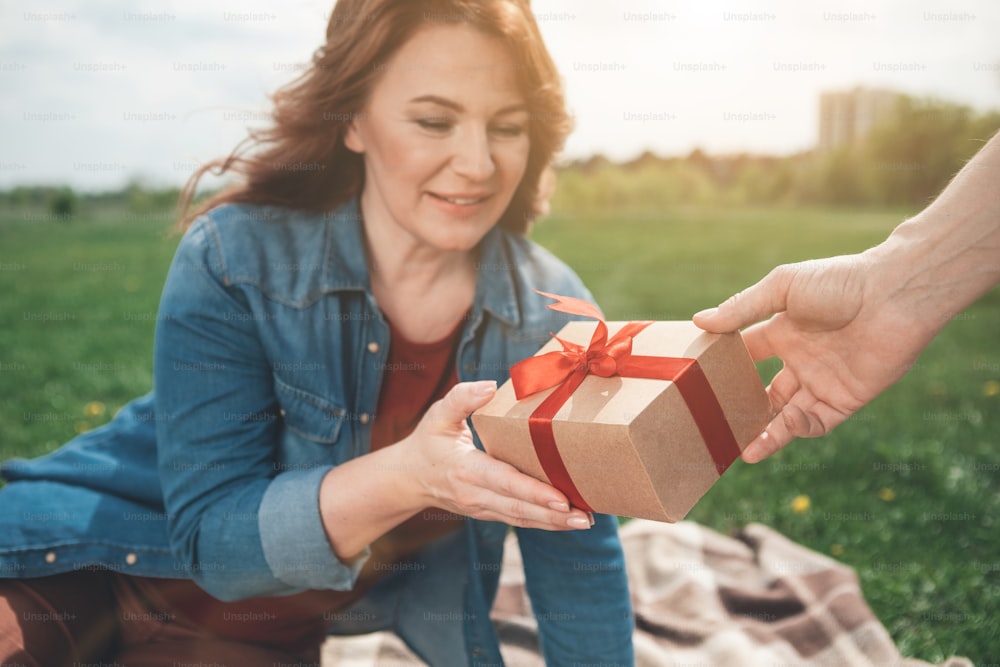 It is for you. Surprised lady is receiving present from her husband. She is looking at it and smiling while relaxing on meadow. Focus on  wrapped box with ribbon in male hand