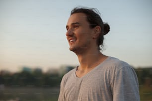Perfil do homem bonito sorrindo mostrando dentes no parque, no lago enquanto olhava para o nascer do sol. Retrato do homem atlético positivo da capoeira no fundo da praia da cidade, vestindo camiseta cinza.