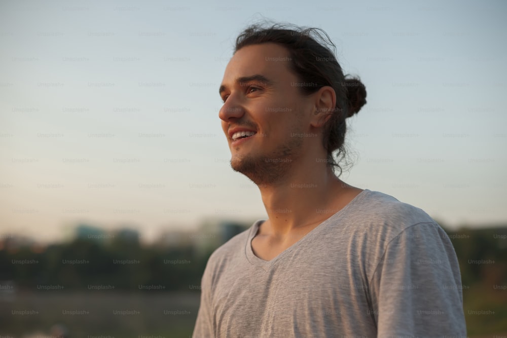 Profil eines gutaussehenden Mannes, der lächelt und Zähne im Park zeigt, auf dem See, während er den Sonnenaufgang betrachtet. Porträt des sportlich positiven Capoeira-Mannes auf Stadtstrandhintergrund, der ein graues T-Shirt trägt.