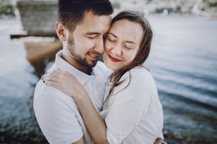 happy stylish couple in love hugging on the beach in  summer city. modern woman and man in fashionable white clothes embracing at the water, romantic sensual moment