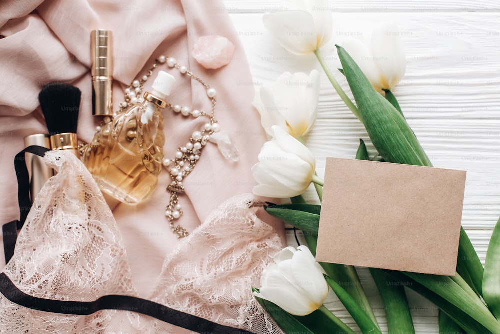 Bijoux de lingerie femme et parfum sur tissu doux et tulipes avec carte de voeux vide sur fond rustique blanc. Des essentiels pour les filles pour des vacances. Préparation de mariage bohème le matin