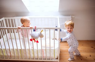 Two happy toddler children in bedroom at home. A girl in a cot and a boy standing on the floor.
