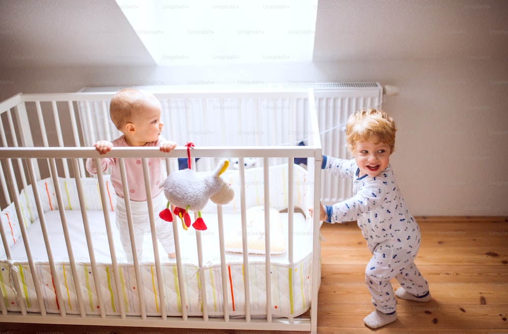 Two happy toddler children in bedroom at home. A girl in a cot and a boy standing on the floor.