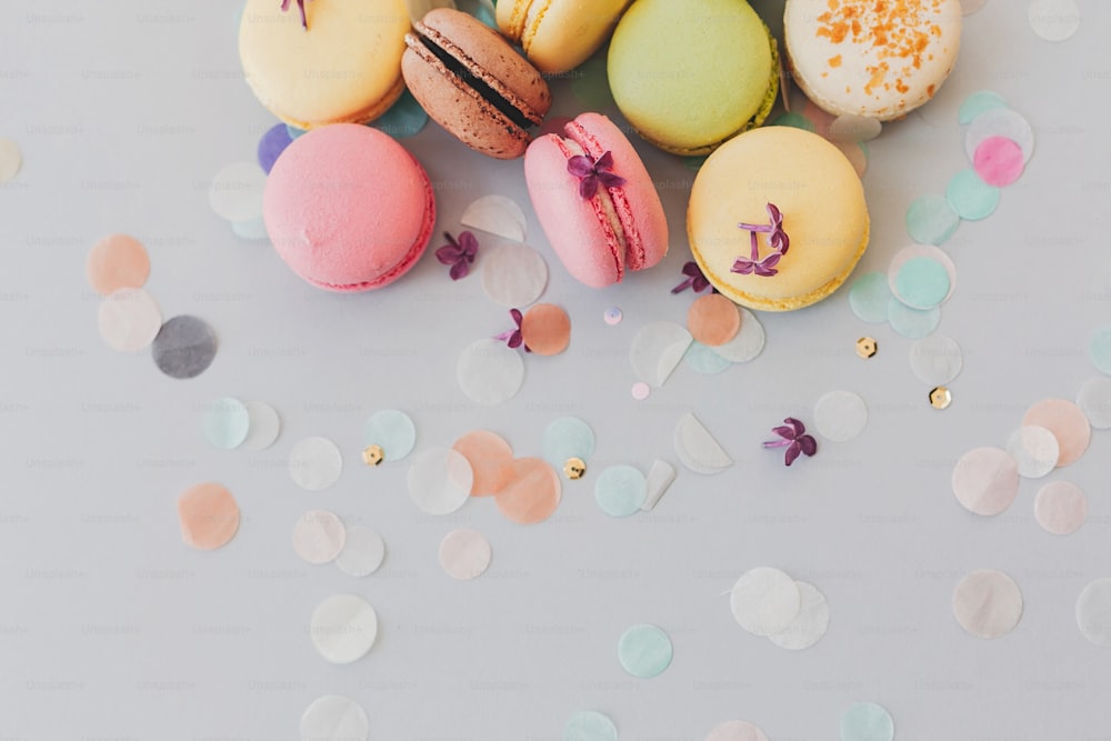delicious colorful macaroons on trendy pastel gray paper with lilac flowers and confetti, flat lay. tasty pink, yellow, green and brown macaroons. space for text. candy for party