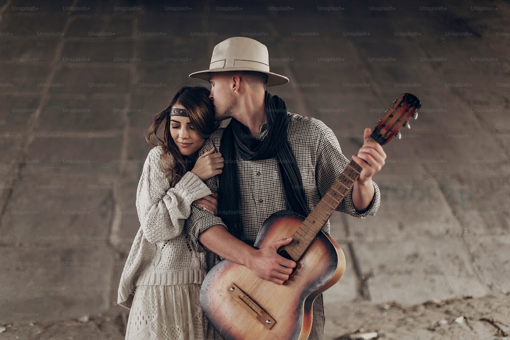 Stilvolles Hipster-Paar umarmt sich sanft. Mann mit Hut spielt Gitarre für seine Boho-Frau im Strickpullover. atmosphärischer sinnlicher Moment. rustikaler modischer Look.