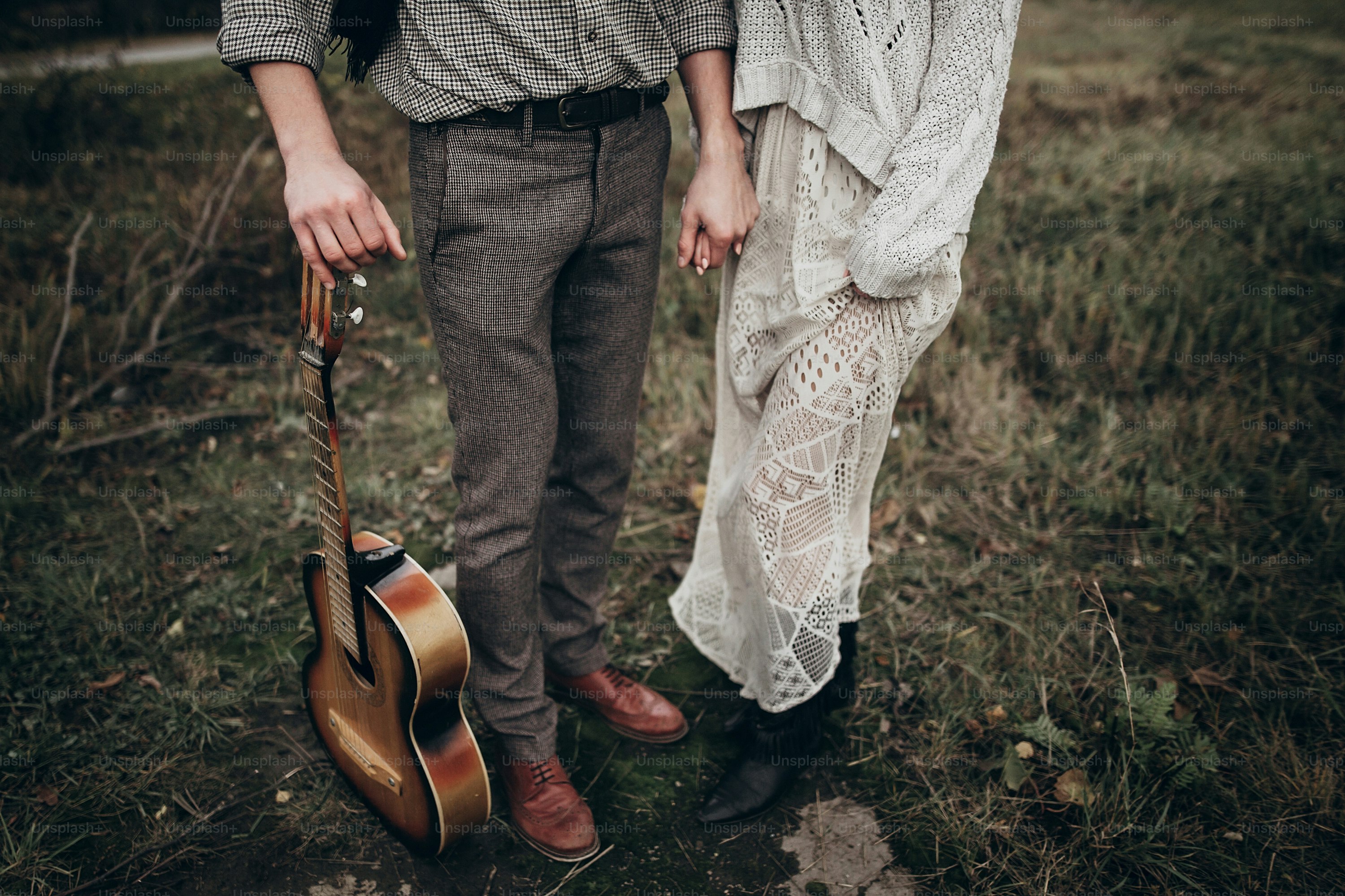 Stilvolles Hipster-Paar, das sich im windigen Feld an den Händen hält. Boho Zigeunerin Frau und Mann mit Gitarre umarmt. atmosphärischer Moment. Modischer Look. Rustikales Hochzeitskonzept