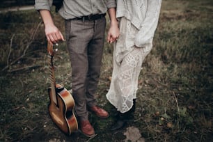 stylish hipster couple holding hands closeup in windy field. boho gypsy woman and man with guitar  hugging. atmospheric moment. fashionable look. rustic wedding concept
