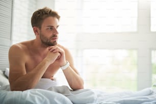 Side view of pensive young male sitting on bed in morning. He is contemplating on day plans before getting up surrounded by white linens. Copy space in right side