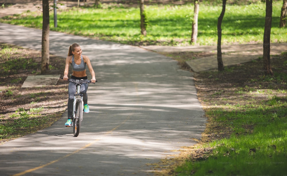 La dama bonita se siente feliz mientras anda en bicicleta por el parque a lo largo de un carril ancho. Ella está sonriendo mientras pedalea y examina el entorno que la rodea. Copiar espacio en el lado derecho