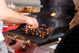 man roasting mushrooms, holding tongs, on open grill, outdoor kitchen. chef making vegetable meal, food festival in city. tasty food, food-court. summer picnic