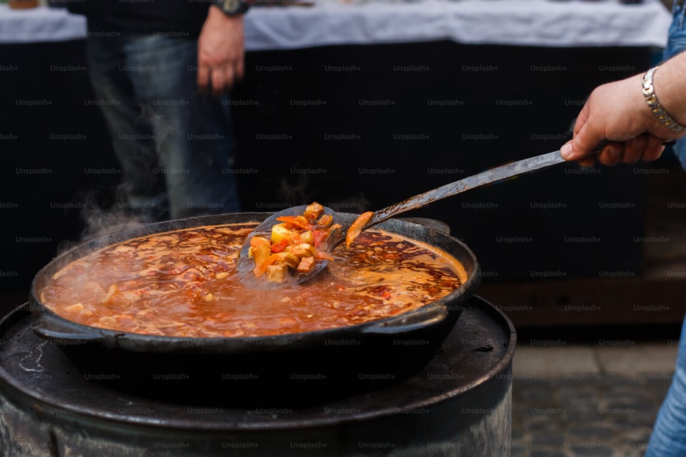 Delicioso goulash en tazón grande con cuchara en parrilla abierta, cocina al aire libre. Chef que prepara deliciosa comida tradicional húngara, festival gastronómico en la ciudad. Comida sabrosa, patio de comidas. Picnic de verano
