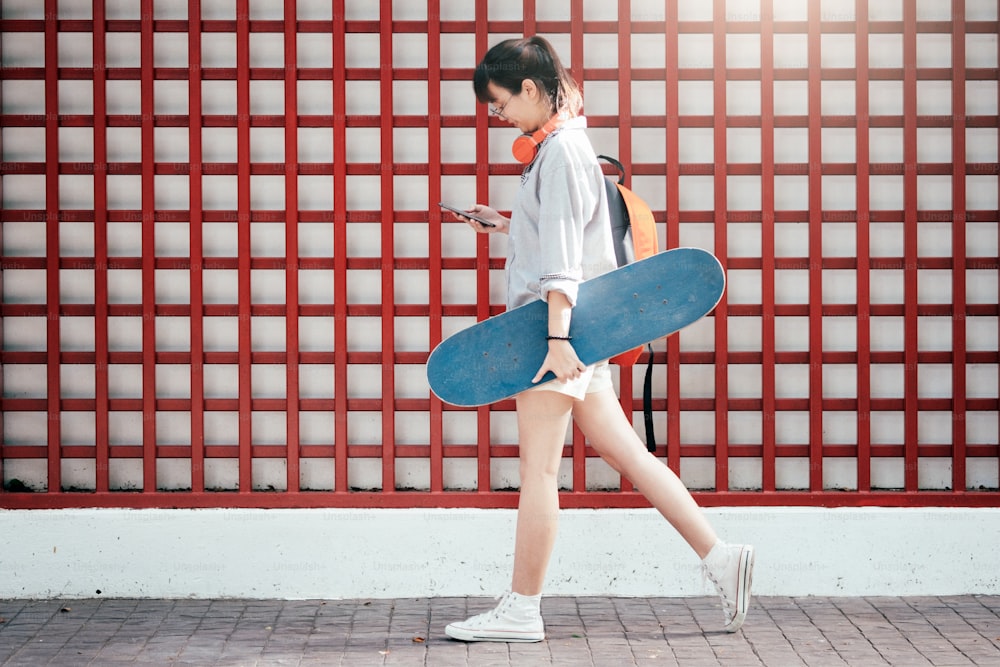 Giovane ragazza asiatica hipster che cammina e tiene lo skateboard e usa il telefono cellulare per chattare.