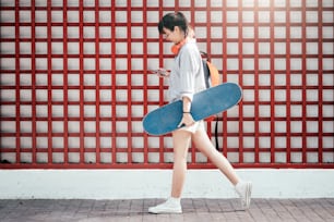 Young hipster asian girl walking and holding skateboard and using mobile phone to chating.