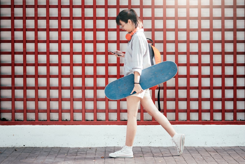 Young hipster asian girl walking and holding skateboard and using mobile phone to chating.