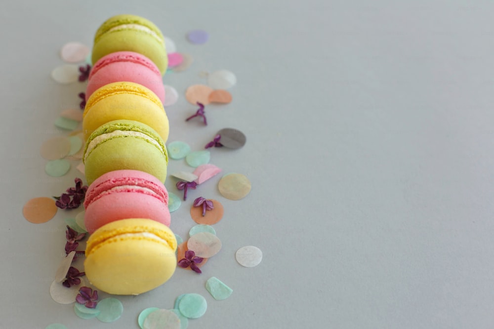 Bunte Macarons auf trendigem pastellgrauem Papier mit lila Blüten und Konfetti. leckere rosa, gelbe, grüne und braune Makronen. Schokoriegel für Party. Food-Fotografie. leckerer Hintergrund