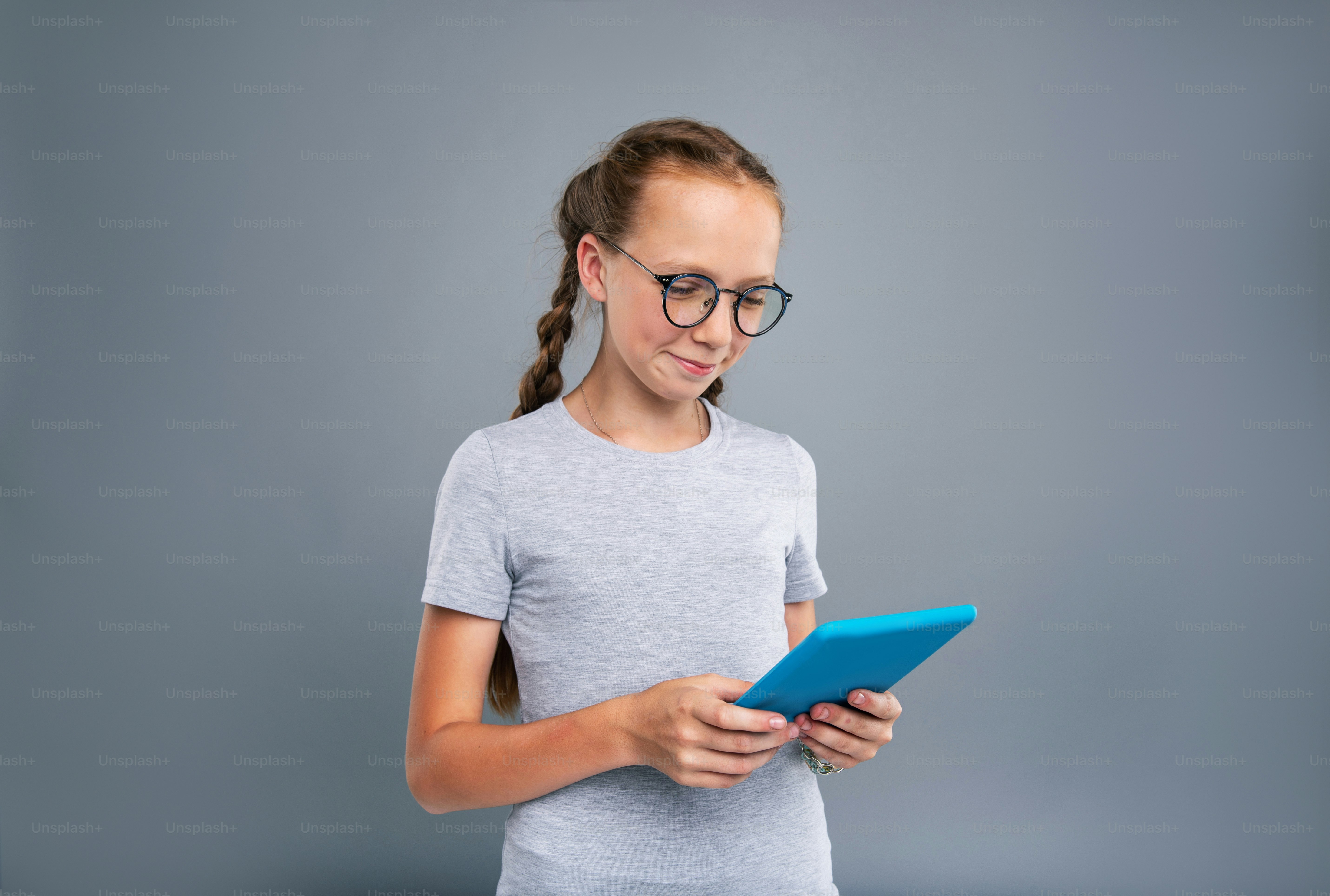 Cute nerd. Cheerful teenage girl in eyeglasses reading from a blue tablet and smiling while standing isolated on a blue-grey background