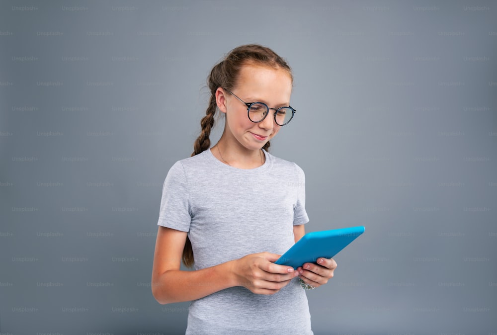 Süßer Nerd. Fröhliches Teenagermädchen mit Brille, das von einem blauen Tablet liest und lächelt, während es isoliert auf einem blaugrauen Hintergrund steht