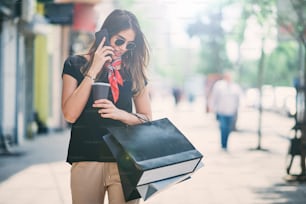 Retrato de una mujer sosteniendo bolsas de papel y café en la calle después de shoping mientras usa un teléfono inteligente.