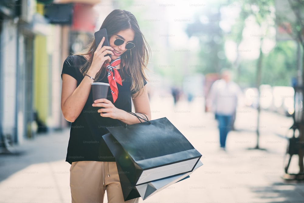 Portrait of woman holding paper bags and coffee on the street after shoping while using smart phone.