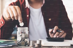 Homme mettant de l’argent (pièce de monnaie) dans le bocal en verre pendant l’utilisation calculatrice pour calculer le revenu ou l’impôt - Financial Business Concept