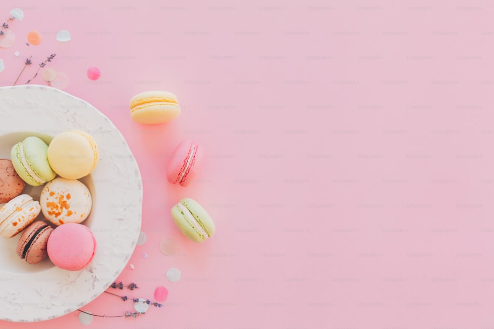 Délicieux macarons colorés dans une assiette blanche vintage sur papier rose pastel à la mode, pose à plat élégante. Savoureux macarons roses, jaunes, verts et bruns à la lavande et aux confettis. espace pour le texte