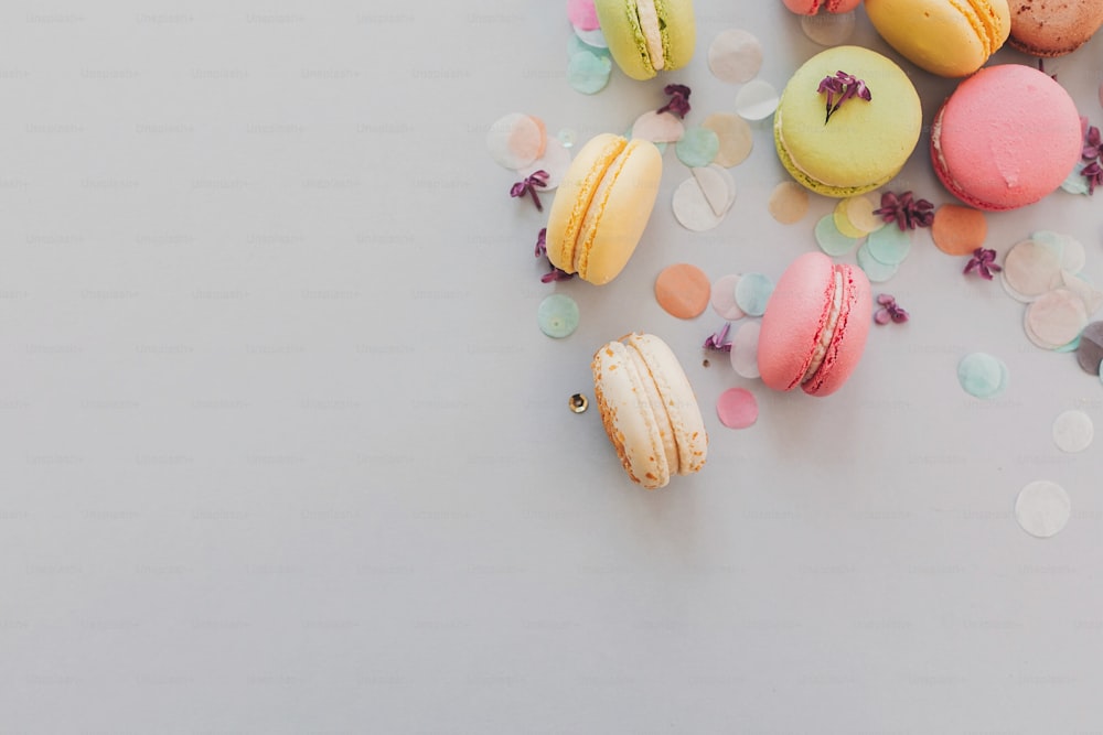 Délicieux macarons colorés sur papier gris pastel tendance avec des fleurs lilas et des confettis, pose à plat. Savoureux macarons roses, jaunes, verts et bruns. espace pour le texte. bonbons pour la fête