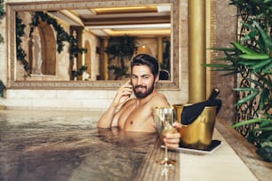 Young handsome bearded man enjoying and relaxing in exclusive spa resort. He sitting in hot tub and drinking wine.