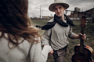 man in hat with guitar looking and smiling to his boho woman in windy field. atmospheric sensual moment. fashionable look. rustic wedding concept.
