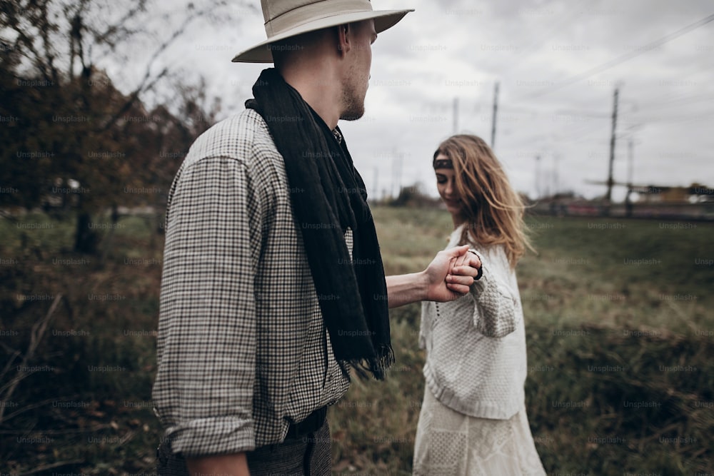 stylish hipster couple holding sensually hands. boho gypsy woman and man in hat embracing in windy field. atmospheric amazing moment. fashionable look. rustic wedding concept