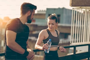 Young couple standing on a building rooftop terrace, drinking water and speaking while taking a break from a workout
