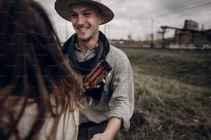man in hat with guitar looking and smiling to his boho woman in windy field. atmospheric sensual moment. fashionable look. rustic wedding concept.
