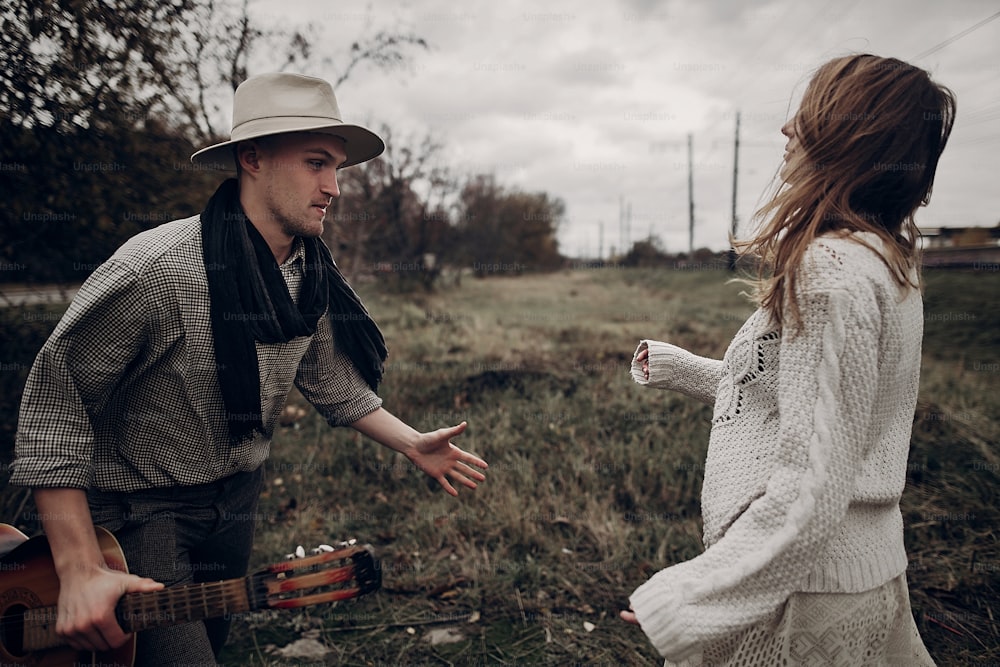 Couple hipster romantique, bel homme musicien avec guitare et femme gitane en pull bohème dansant dans le champ d’automne