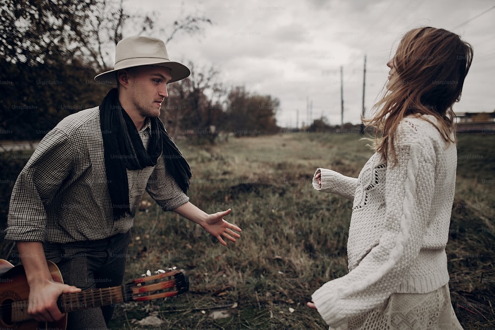 Coppia hipster romantica, bell'uomo musicista con la chitarra e donna zingara in maglione boho che balla nel campo autunnale