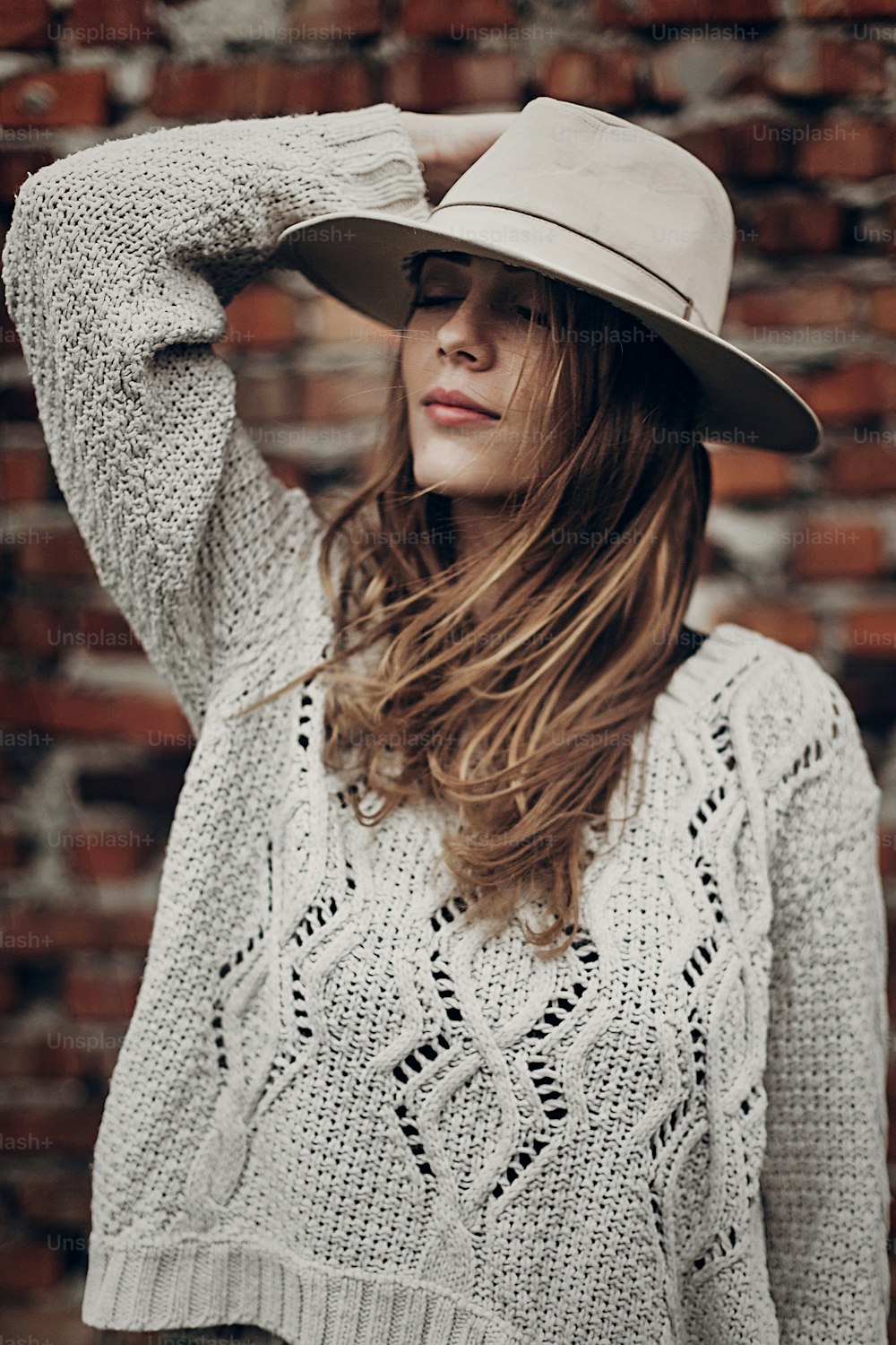 Stilvolle Hipster-Frau, die in einem Strickpullover auf dem Hintergrund einer Ziegelmauer posiert und einen Hut hält. atmosphärischer sinnlicher Moment. Boho Country modischer Look. Freie Menschen